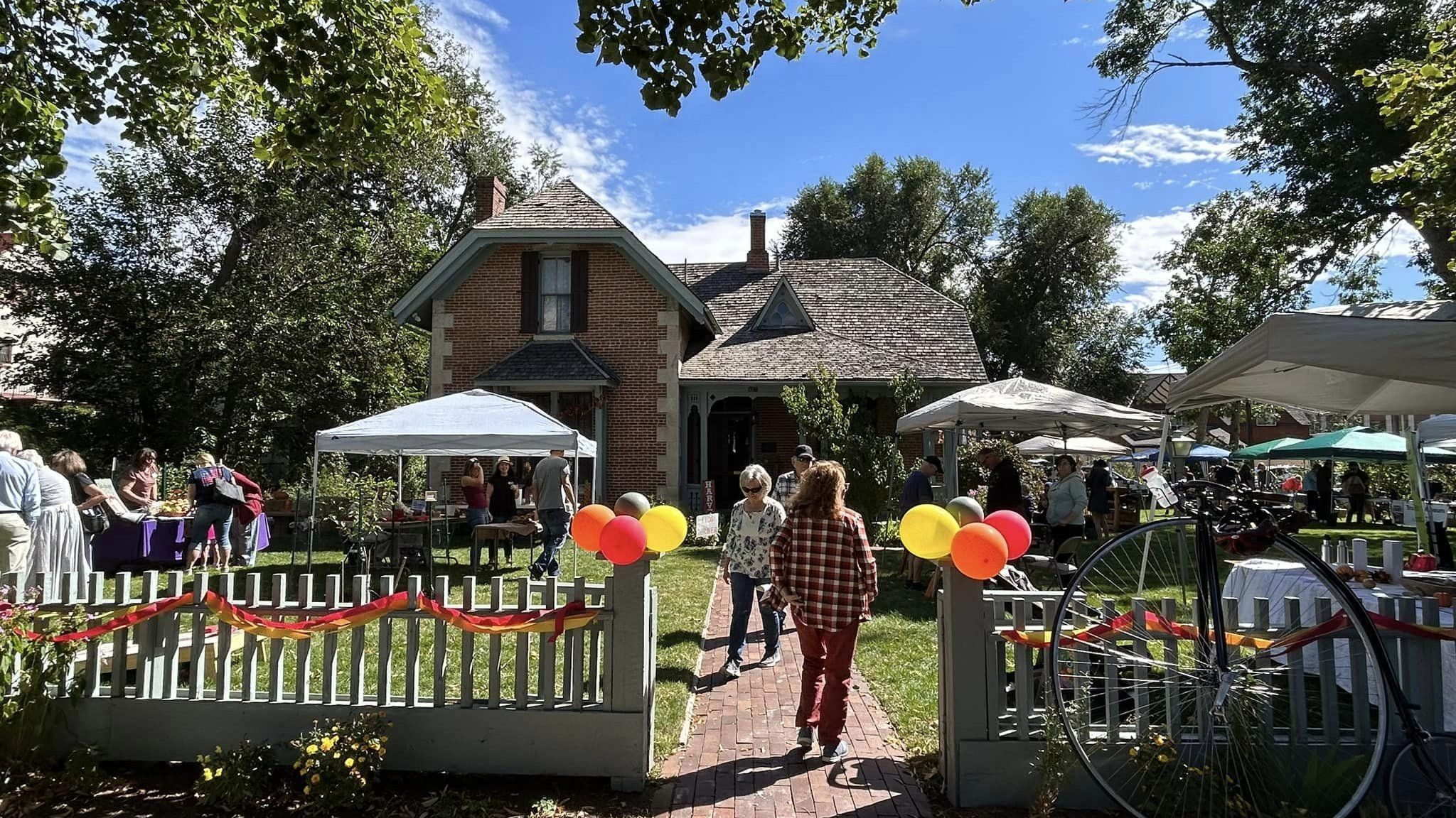 McAllister House during the Harvest Festival featuring colorful balloons, a lively front walkway leading to the house, people gathered at the event, pop-up vendor tents, and lush trees under a clear blue sky.
